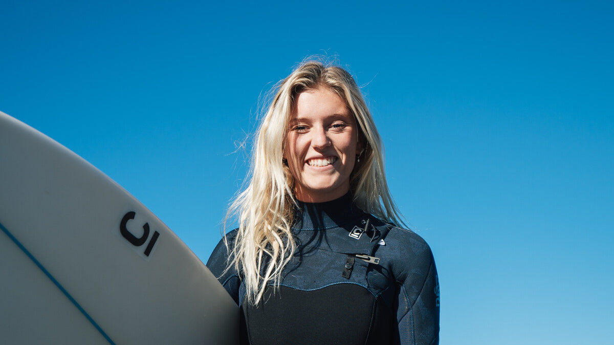 Sophie Fletcher smiling at the camera wearing a wetsuit and holding surfboard. blonde hair, blue skies.