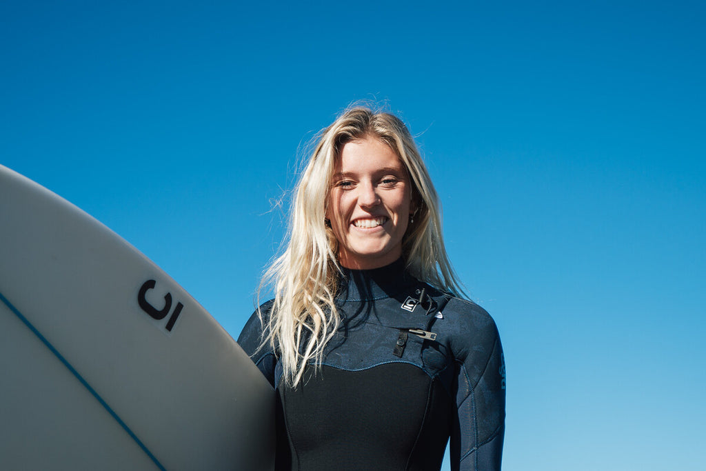Sophie Fletcher smiling at the camera wearing a wetsuit and holding surfboard. blonde hair, blue skies.